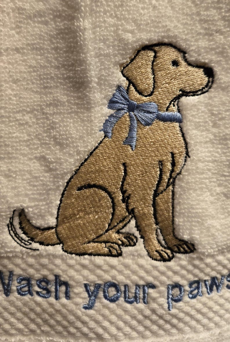 May include: Embroidered white towel featuring a tan dog with a blue bow. The dog is sitting, with the text "Wash your paws" embroidered in blue below. The towel has a textured surface.