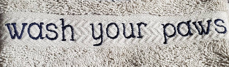 May include: A light gray towel with the embroidered text "wash your paws" in navy blue. The text is centered on a textured band with a chevron pattern.
