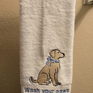 May include: White hand towel with an embroidered tan dog wearing a blue bow. The text "Wash your paws" is embroidered below the dog. The towel is hanging on a silver towel bar.