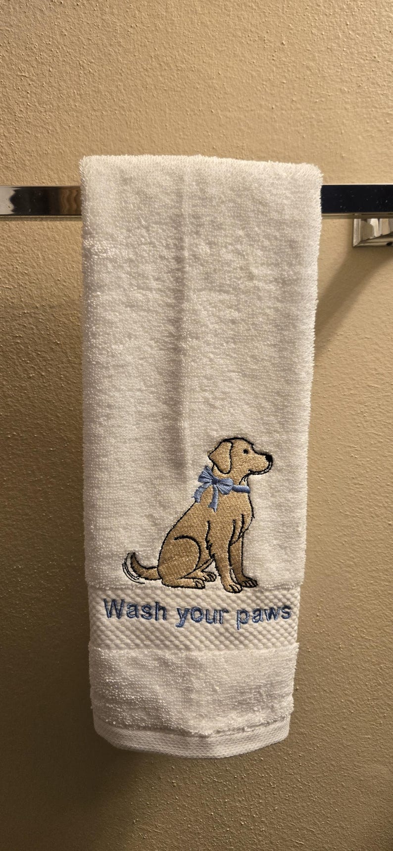 May include: White hand towel with an embroidered tan dog wearing a blue bow. The text "Wash your paws" is embroidered below the dog. The towel is hanging on a silver towel bar.