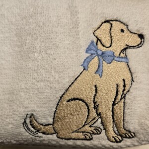 May include: White hand towel with an embroidered image of a golden Labrador Retriever wearing a light blue bow. The dog is sitting, facing right, with black outline details.