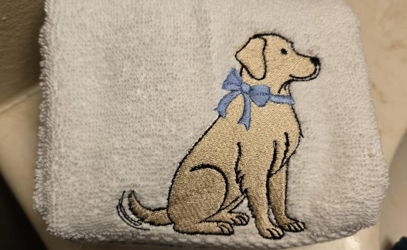 May include: White hand towel with an embroidered image of a golden Labrador Retriever wearing a light blue bow. The dog is sitting, facing right, with black outline details.