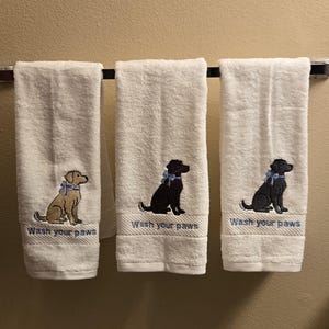 May include: Three white hand towels with embroidered dog designs and the text "Wash your paws". The dogs are in shades of brown and black, each wearing a blue bow. The towels are hanging on a chrome towel bar.