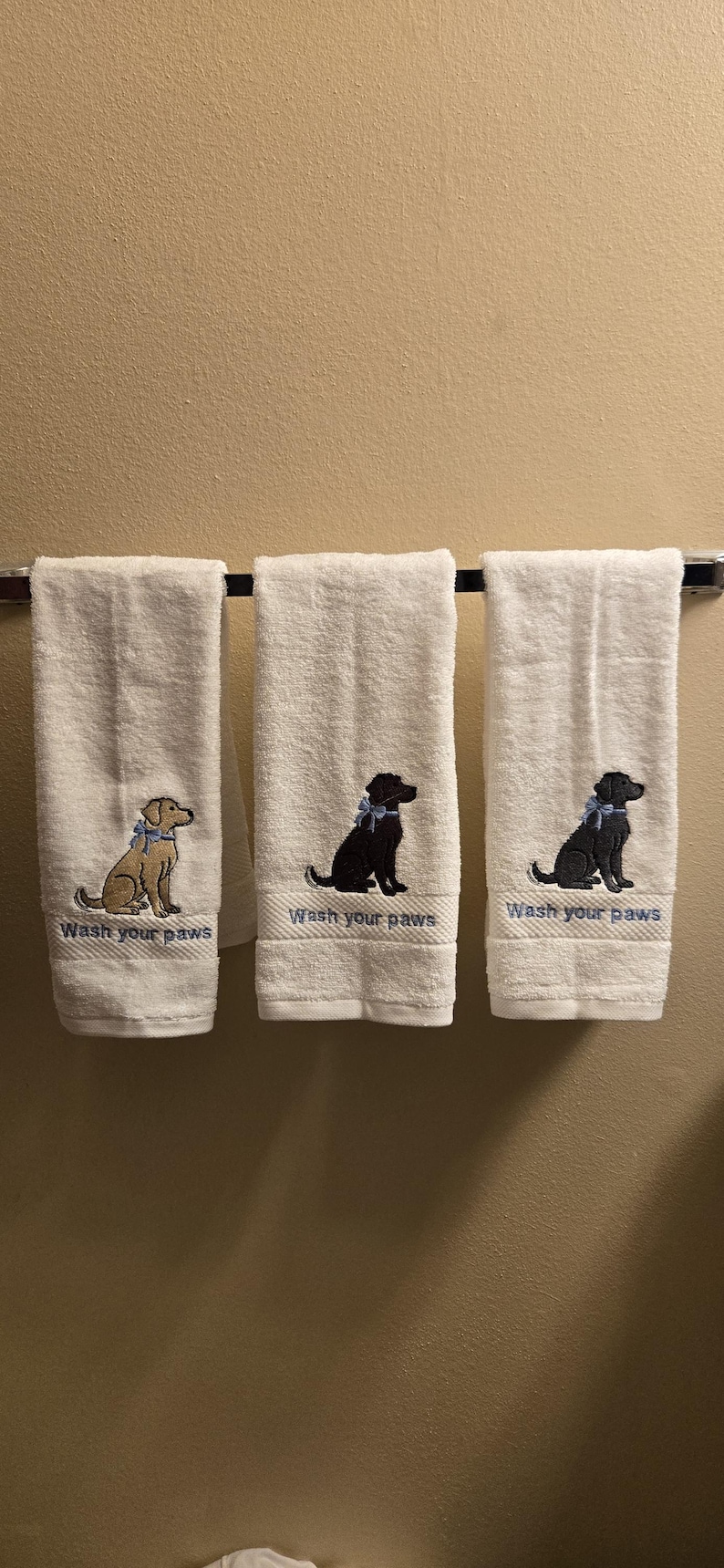May include: Three white hand towels with embroidered dog designs and the text "Wash your paws". The dogs are in shades of brown and black, each wearing a blue bow. The towels are hanging on a chrome towel bar.