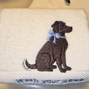 May include: White hand towel with an embroidered brown dog wearing a blue bow. The towel has the words "Wash your paws" embroidered in blue at the bottom. The towel is made of a soft, absorbent material.