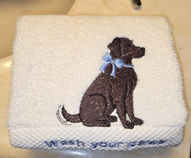 May include: White hand towel with an embroidered brown dog wearing a blue bow. The towel has the words "Wash your paws" embroidered in blue at the bottom. The towel is made of a soft, absorbent material.