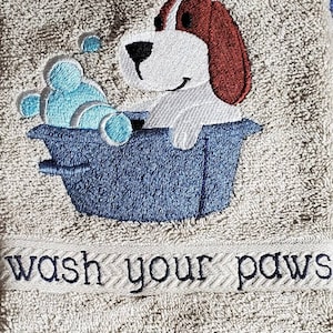 May include: A beige bath towel with an embroidered design of a cartoon dog in a blue tub with blue bubbles. The text "wash your paws" is embroidered below the dog.
