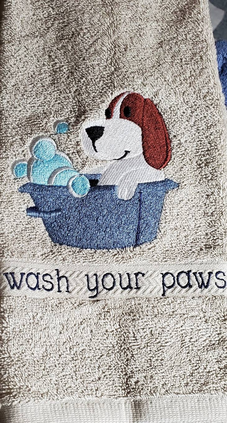 May include: A beige bath towel with an embroidered design of a cartoon dog in a blue tub with blue bubbles. The text "wash your paws" is embroidered below the dog.