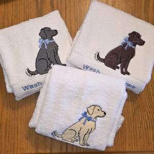 May include: Three white washcloths with embroidered dog designs. Each cloth features a different colored dog: gray, brown, and tan, all wearing a blue bow. The word "Wash" is embroidered in blue on each cloth.