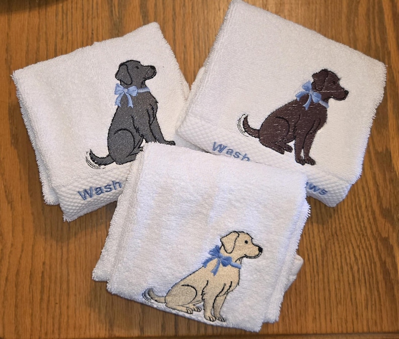 May include: Three white washcloths with embroidered dog designs. Each cloth features a different colored dog: gray, brown, and tan, all wearing a blue bow. The word "Wash" is embroidered in blue on each cloth.