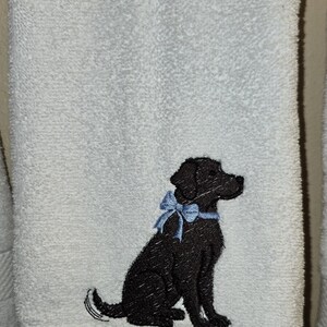 May include: White bath towel with an embroidered black Labrador Retriever wearing a blue bow. The text "Wash your paws" is embroidered in blue below the dog. The towel is hanging on a bar.