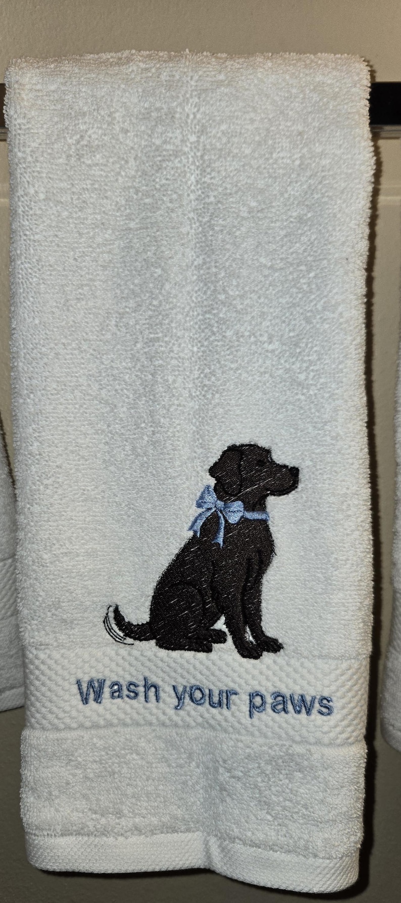 May include: White bath towel with an embroidered black Labrador Retriever wearing a blue bow. The text "Wash your paws" is embroidered in blue below the dog. The towel is hanging on a bar.