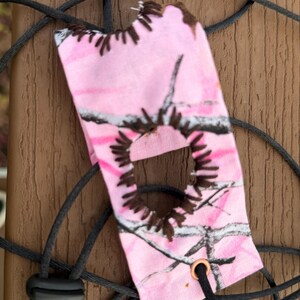 Puede incluir: Una bolsa de tela rectangular con un patrón de camuflaje rosa y blanco, que presenta un recorte circular central bordeado con costuras de color marrón oscuro. Incluye un cordón negro con un cierre de plástico y un ojal de metal color cobre.