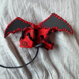 May include: A red fabric accessory featuring a black skull and crossbones print, accented with black bat-shaped wings. The edges are finished with thick red stitching, and the piece includes black eyelets with a black cord and a plastic toggle.