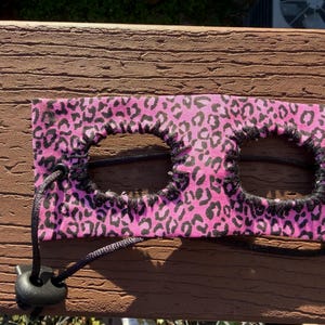 May include: A rectangular pink fabric accessory featuring a black leopard print pattern. The item has two circular cutouts edged with black stitching and includes a black adjustable cord with a plastic toggle for securing the fit around the head.