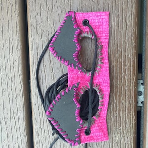 Puede incluir: Un antifaz con una base de tela rosa brillante con un patrón texturizado. Dos parches oculares angulares de color gris oscuro están unidos al frente con puntadas de festón en rosa vibrante. Incluye ojales negros y cordones negros.