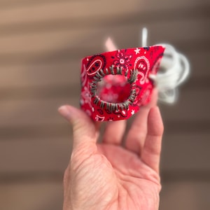 Puede incluir: Un accesorio de tela roja con un patrón de cachemir y estrellas blancas. El artículo tiene una abertura circular reforzada con bordado gris oscuro hecho a mano y cuenta con cordones blancos que sobresalen de un lado.