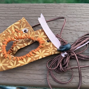 Puede incluir: Un accesorio de tela color mostaza con un patrón repetitivo de margaritas amarillo claro y un recorte central bordeado con hilo de bordar naranja grueso. Incluye un cordón largo marrón con un tope oscuro y una etiqueta de cinta rosa pálido.