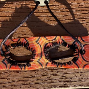 May include: A rectangular fabric mask featuring an orange and yellow circular pattern with two oval eye openings edged in black thread. A black cord with a plastic toggle passes through two black metal grommets at the top corners of the mask.