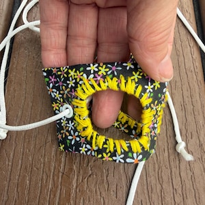 May include: A black fabric accessory featuring a colorful floral pattern of small daisies in white, yellow, pink, and blue. The center hole is reinforced with thick, bright yellow embroidery stitching, and a white cord is threaded through a metal eyelet.