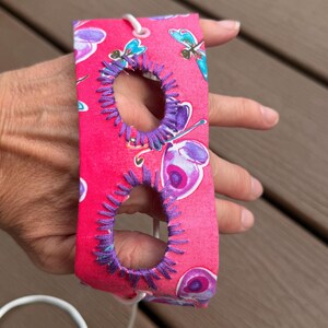 Puede incluir: Una correa rectangular de tela rosa con un patrón repetitivo de mariposas y libélulas coloridas. La correa incluye dos aberturas ovaladas reforzadas con un grueso bordado morado y un cordón blanco enhebrado a través de los extremos.