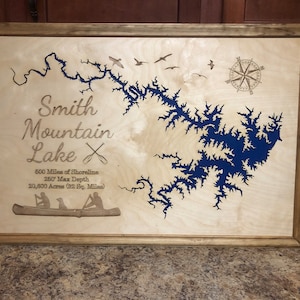 May include: A wooden plaque with a blue map of Smith Mountain Lake. The plaque includes the lake's name, a compass, and details about the lake, including its shoreline length, maximum depth, and acreage.