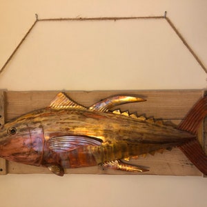 May include: A copper-colored metal fish sculpture mounted on a wooden plank with a rope hanger. The fish has a detailed design and a realistic appearance.