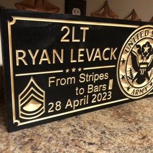 May include: A black wooden plaque with gold lettering and a gold United States Army seal. The plaque reads "2LT RYAN LEVACK From Stripes to Bars 28 April 2023".