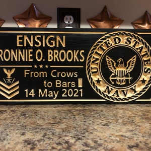 May include: A black wooden plaque with gold lettering and a carved United States Navy emblem. The plaque reads "ENSIGN RONNIE O. BROOKS From Crows to Bars 14 May 2021".