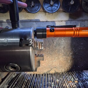May include: An orange flashlight with a black clip and the word "Wurkkow" printed on the side. The flashlight is mounted in a metal chuck.
