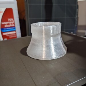 May include: A clear plastic 3D printed funnel with a wide base and a narrow opening. The funnel is sitting on a gray surface.