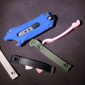 May include: A set of five tools, including a blue utility knife, a pink plastic tool, a white plastic tool, a black plastic tool, and a green plastic tool. The tools are all on a dark gray surface.