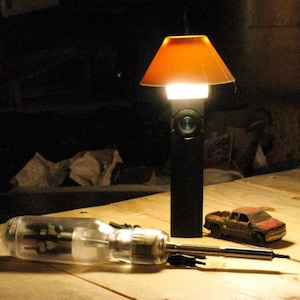 May include: A black rectangular flashlight with a glowing orange conical lampshade attached to the top. Next to the flashlight lies a clear plastic screwdriver with a metal shaft and a small, weathered, red and brown toy pickup truck.