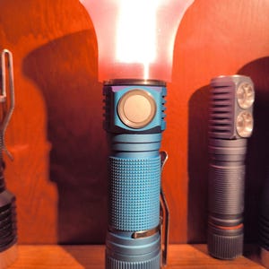 May include: A blue flashlight with a textured grip and a bright, glowing red diffuser. The flashlight has a metal clip and a large button. Other flashlights are visible in the background.