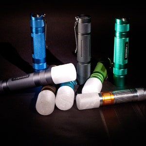 May include: A group of five flashlights in various colors, including blue, green, and gray, with white diffusers. The flashlights are arranged in a semi-circle on a black surface.
