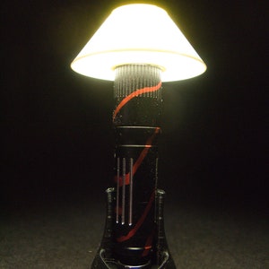 May include: A small, decorative lamp with a black base and a white lampshade. The lamp's body is black with red accents. The lampshade is conical and emits a warm, yellow light, creating a cozy ambiance. The lamp is designed for a desk or bedside table.