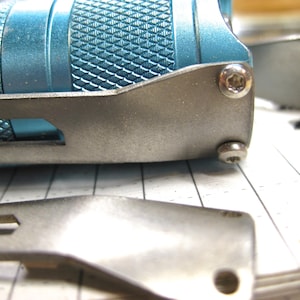 May include: Close-up of a multi-tool with a textured blue handle and silver metal components. The tool is partially disassembled, with a blade and other parts visible. The image is on a lined paper surface.