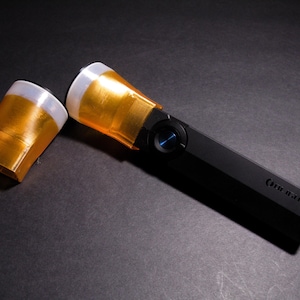 May include: A black flashlight with a gold-colored head and a blue button. The flashlight has a rectangular body and a separate gold-colored head cover. The brand name is printed on the side.
