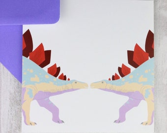 May include: A white card with two cartoon dinosaurs facing each other. The dinosaurs are light blue, purple, and cream colored with red spines. The card is blank for writing a message.