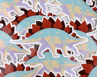 Puede incluir: Una pila de pegatinas de dinosaurios coloridos con un patrón azul, amarillo y morado. Los dinosaurios tienen una espalda espinosa y están sobre un fondo rojo.