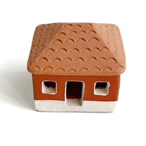 May include: A small house-shaped figurine made of terracotta with a textured, scalloped roof. The structure features a reddish-brown upper section and a white base, with two square windows and a central rectangular doorway opening on the front.