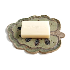May include: A rectangular off-white bar of soap with the embossed text freshwater AUSTRALIA resting on a flat, leaf-shaped ceramic dish. The dish features a speckled light green glaze with dark brown, seed-like indentations across the surface.