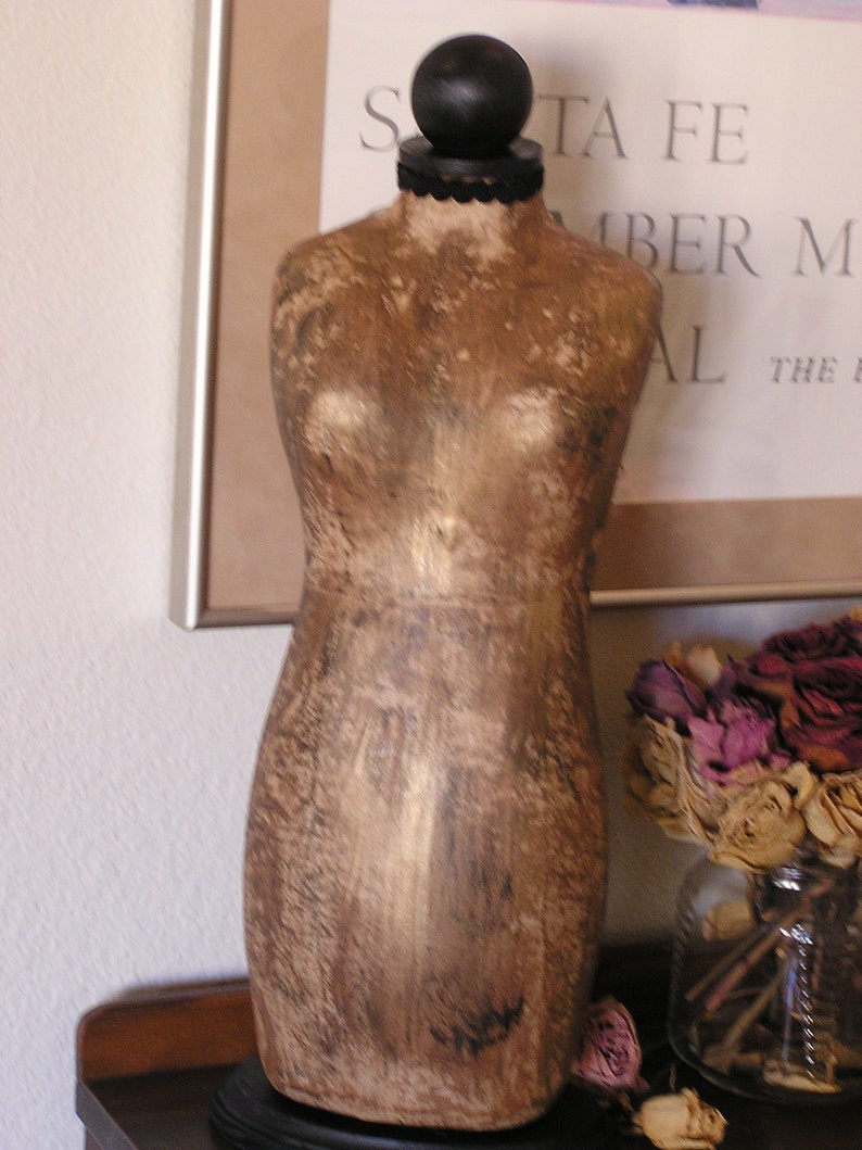 May include: A vintage-style dressmaker's mannequin in a warm, metallic gold finish. The torso is mounted on a black base, with a black ball at the top. The mannequin is displayed near a framed print and a vase of dried flowers.