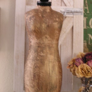 May include: A vintage-style, gold-toned dress form with a black base and a black ball at the top. The dress form is set against a white wooden door with a rustic aesthetic. Dried roses in a glass vase are in the background.