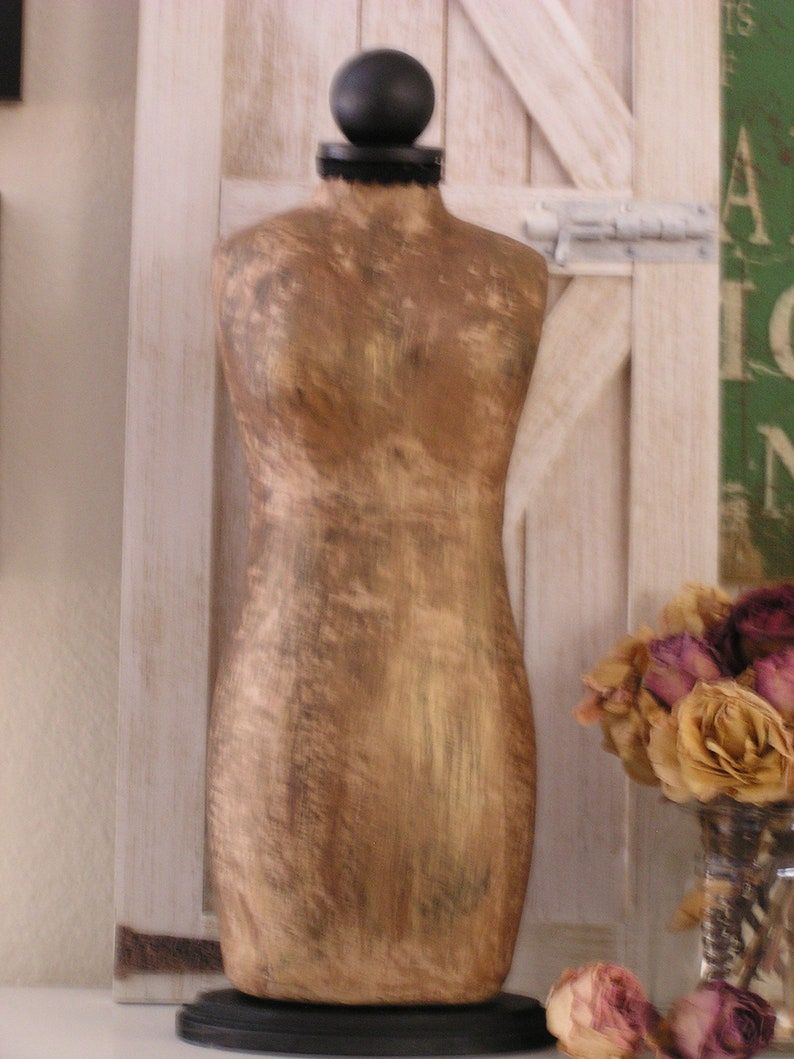 May include: A vintage-style, gold-toned dress form with a black base and a black ball at the top. The dress form is set against a white wooden door with a rustic aesthetic. Dried roses in a glass vase are in the background.