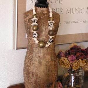 May include: A decorative wooden mannequin torso with a black base and a black ball at the top. A necklace with white, gold, and brown beads is draped around the mannequin. A glass jar with dried roses sits nearby.