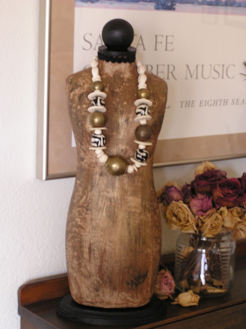May include: A decorative wooden mannequin torso with a black base and a black ball at the top. A necklace with white, gold, and brown beads is draped around the mannequin. A glass jar with dried roses sits nearby.