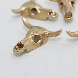 May include: Close-up of several small, gold-colored bull skull charms. Each charm has detailed horns, eye sockets, and a nasal cavity. The charms are arranged on a white surface, suggesting a craft or jewelry-making theme.