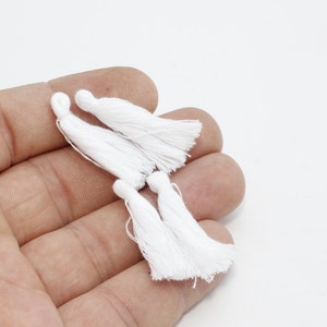 May include: Several small, white tassels are held in a hand against a white background. Each tassel has a looped top and a fringed bottom. The tassels are made of a soft, likely woven material.