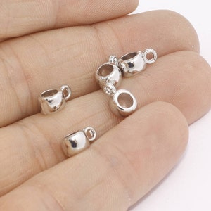 Könnte beinhalten: Eine Sammlung kleiner, glänzender, silberfarbener Tassenanhänger. Jeder Anhänger hat einen abgerundeten Körper mit einem kleinen Henkel an der Seite. Geeignet für Schmuckherstellung oder Bastelprojekte.